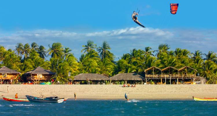 Kitesurfer launching high above palm-fringed beach with thatched huts and fishing boats below