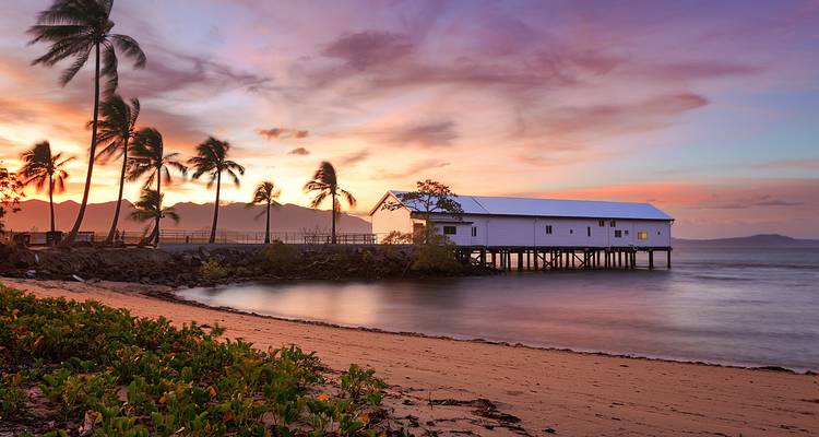 Stilted white boathouse sits over calm tropical waters with palm trees and pastel sunrise colors.