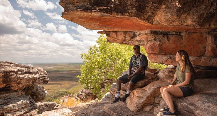 Two hikers rest under a rocky overhang overlooking vast outback plains beneath a bright sky.