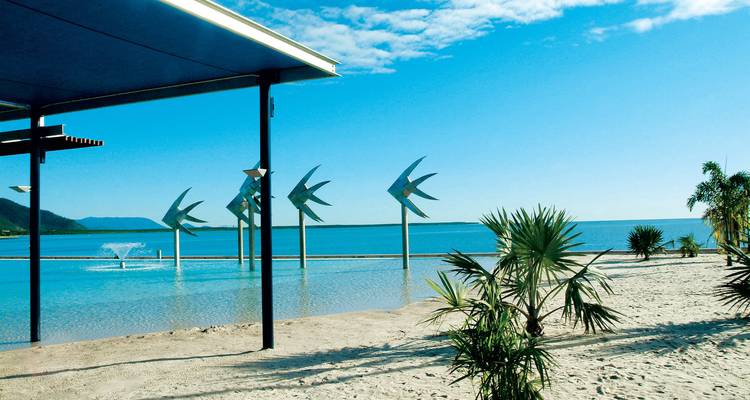 Modern beachfront lagoon with metal fish sculptures overlooking blue sea and distant hills.