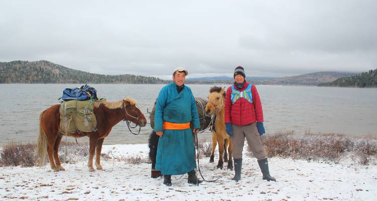 Twee ruiters in traditionele kleding staan met hun lastdieren naast een meer met sneeuwranden.