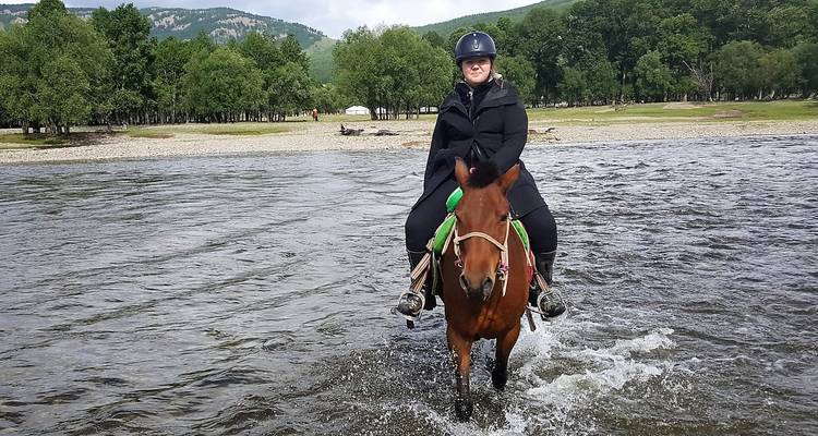 Een ruiter leidt een paard door een ondiep riviertje met spattend water en bosrijke oevers op de achtergrond.