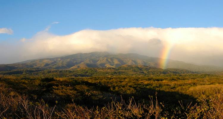 Un arco iris se extiende sobre una cordillera volcánica boscosa bajo un cielo dorado al atardecer.