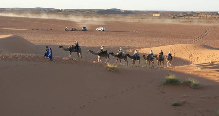 Caravane de touristes chevauchant des chameaux conduits par un guide à travers de hautes dunes de sable orange dans le désert.