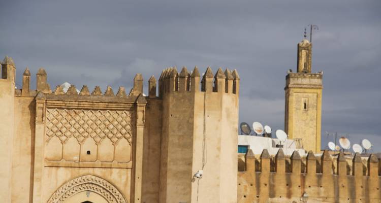 Des murs fortifiés beige avec des créneaux décoratifs et un minaret adjacent surmonté d'antennes satellites sous un ciel gris.