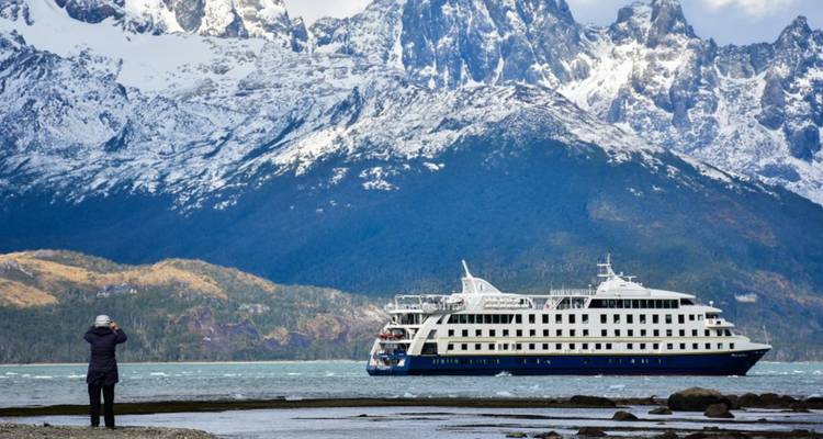 Ein Expeditionskreuzfahrtschiff segelt unter zerklüfteten, schneebedeckten Bergen hindurch, während ein Besucher vom Ufer aus fotografiert.