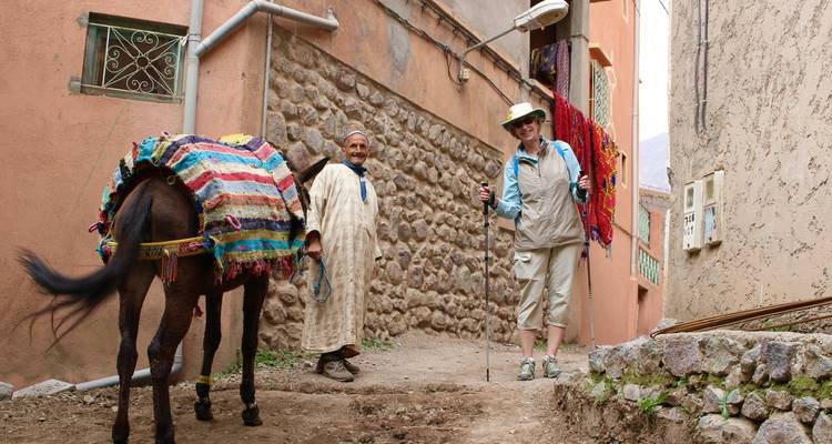 Tourist with trekking poles walks past a donkey and a smiling villager in a narrow village lane.