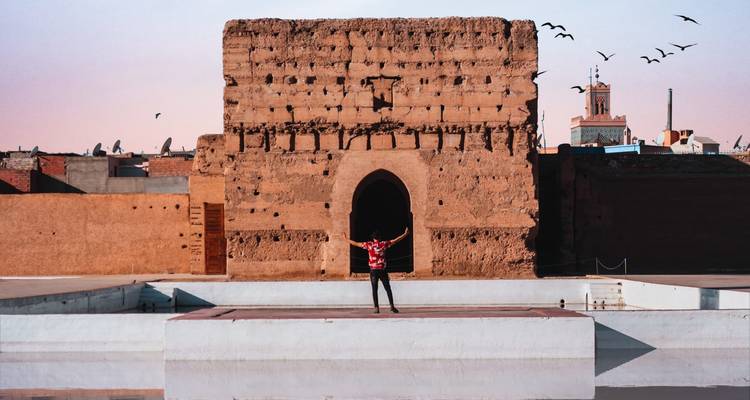 Traveller raising arms in front of an ancient adobe wall at sunset with birds in the sky.