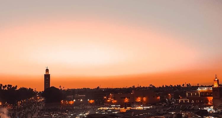 Orange twilight sky over Marrakesh with silhouettes of minarets and bustling market below.