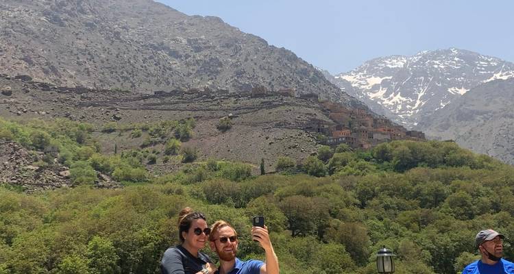 Couple taking a selfie with snow-capped Atlas Mountains and hillside village in the background.