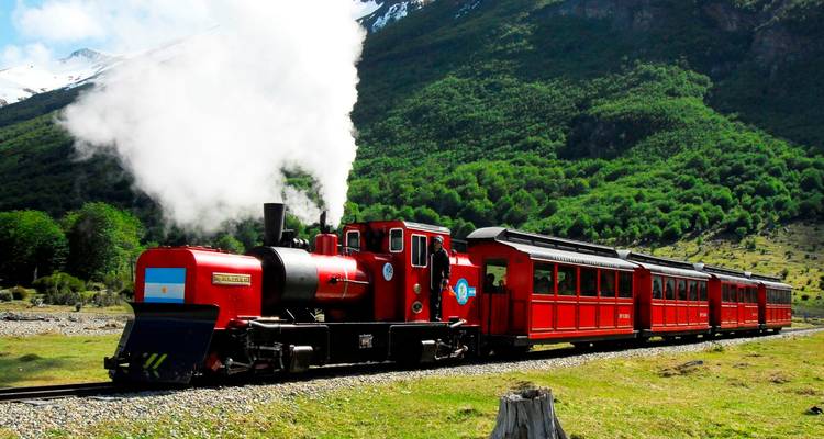 Een felrode stoomtrein puft door groene Patagonische heuvels onder sneeuwwitte bergtoppen.