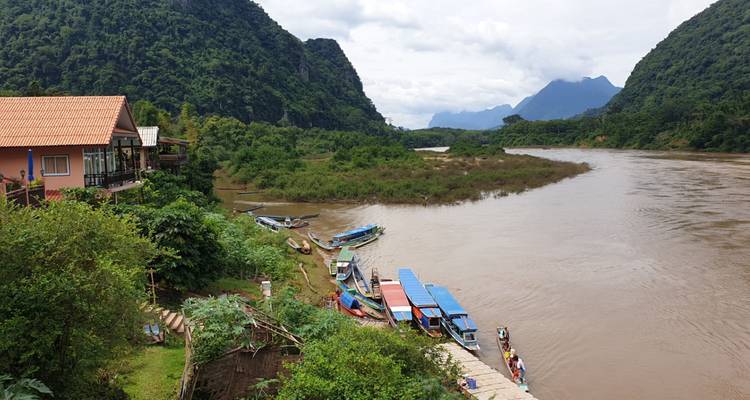 Río amplio con barcas de cola larga amarradas en un pequeño embarcadero de la aldea entre colinas de piedra caliza verde