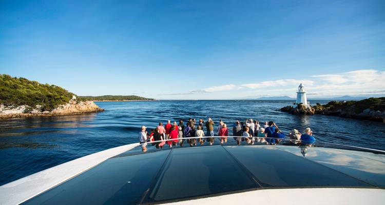 Les passagers se rassemblent à la proue d'un bateau de croisière qui s'approche d'un petit phare blanc à l'embouchure d'une baie.
