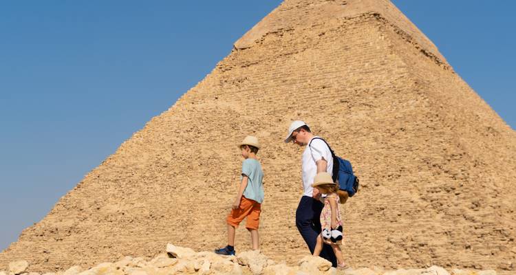 Een vader en twee kinderen wandelen naast een gigantische piramide onder een helder woestijnhemel.