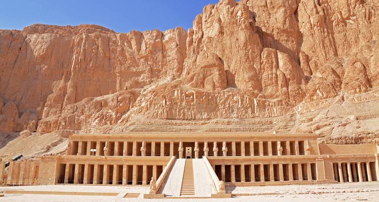 De indrukwekkende voorgevel van de Tempel van Hatshepsut tegen uitstekende steile rotswanden onder een heldere blauwe hemel.