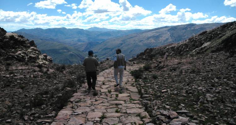 Dos excursionistas caminan por un camino de piedra antiguo a través de las montañas andinas escarpadas bajo un cielo azul.