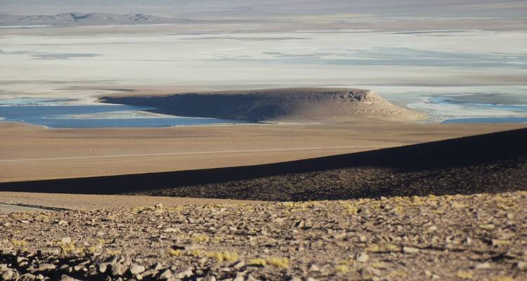 Schaduwrijke woestijnhellingen kijken uit op een vlak zoutpan met hier en daar ondiepe blauwe poelen en een geïsoleerde tafelberg.