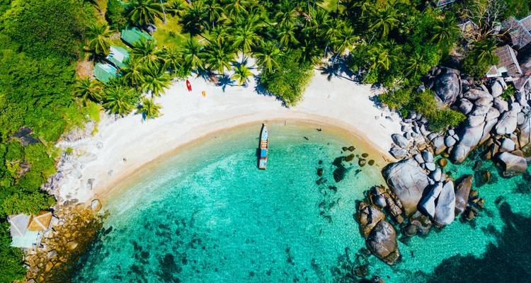 Vista aérea vibrante de una bahía turquesa, playa blanca y palmeras exuberantes con una barca de cola larga anclada.
