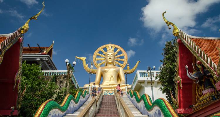 Estatua de la Gran Buda Dorada en la cima de una escalera ornamentada con turistas subiendo bajo un cielo azul.