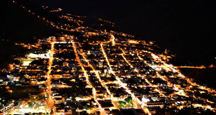 Vista aérea borrosa nocturna de un pueblo de montaña con patrón de cuadrícula iluminado por farolas naranjas.