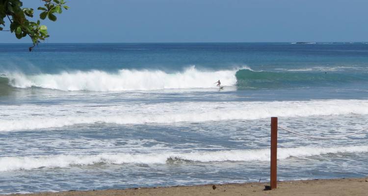 Ein Surfer, der eine brechende Welle auf einem leuchtend blauen Ozean unter klarem Himmel reitet.