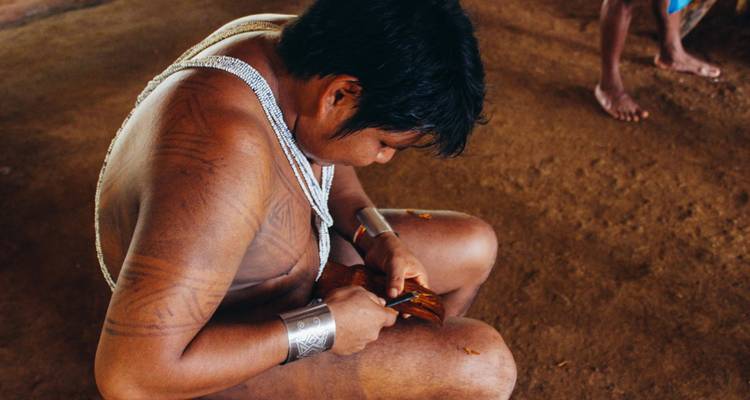 Indigener Mann, der Holz in einer Hütte mit Lehmfußboden und natürlichem Licht bearbeitet.