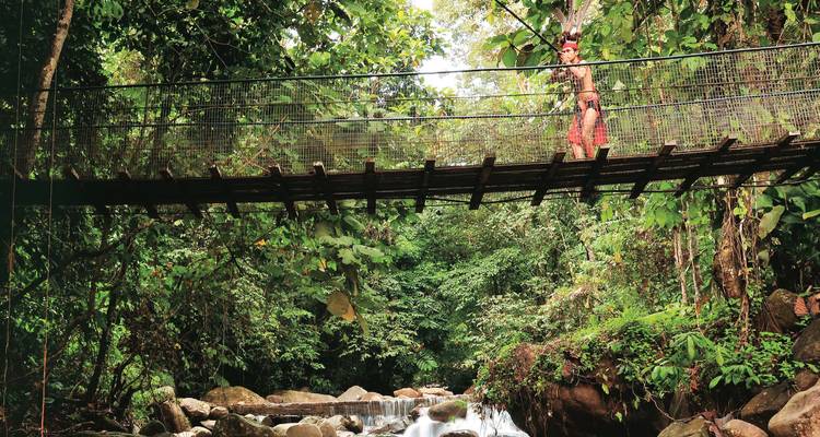 Ein Indigener in traditioneller Kleidung überquert eine schmale Hängebrücke über einen Dschungelbach.