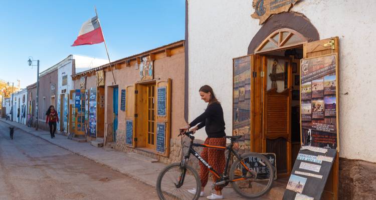 Cycliste et piéton devant des façades de magasins en adobe colorées avec un drapeau chilien flottant en plein jour.