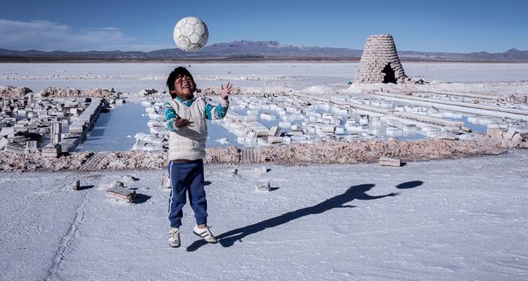Un jeune garçon jetant joyeusement un ballon de football sur les vastes étendues blanches de sel à côté de petites structures en brique.