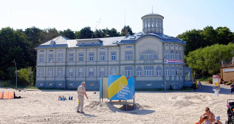 Historic seaside wooden villa with tower windows stands on sandy beach where a man strolls in sunshine.