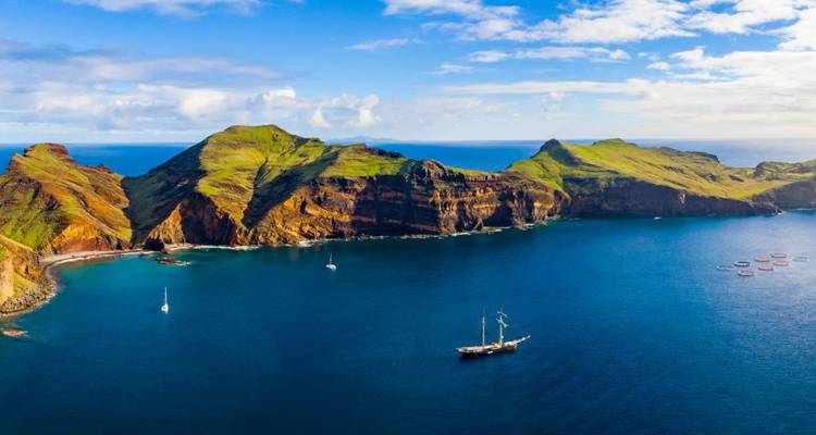 Aerial of verdant Isla de Ons style peninsula with sheer cliffs and anchored sailboats in deep blue Atlantic.