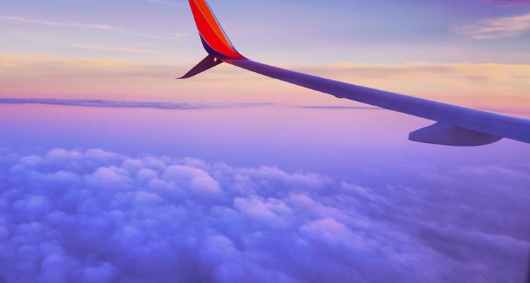 Une aile d'avion tranche au-dessus de nuages pastel au lever du soleil, projetant une douce lueur violette chaude dans le ciel.