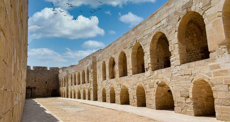 Un long corridor de grès d'une forteresse ancienne bordé d'arches répétées sous un ciel bleu éclatant avec des oiseaux au-dessus.
