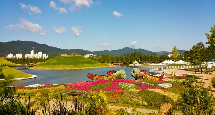 Colorful flower gardens beside a lake with city skyline and forested mountains in the background.