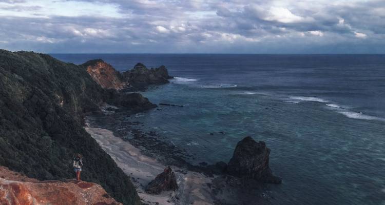 Einsamer Wanderer steht auf einer Klippe und überblickt eine dramatische felsige Küstenlinie und das weite blaue Meer unter düsteren Wolken