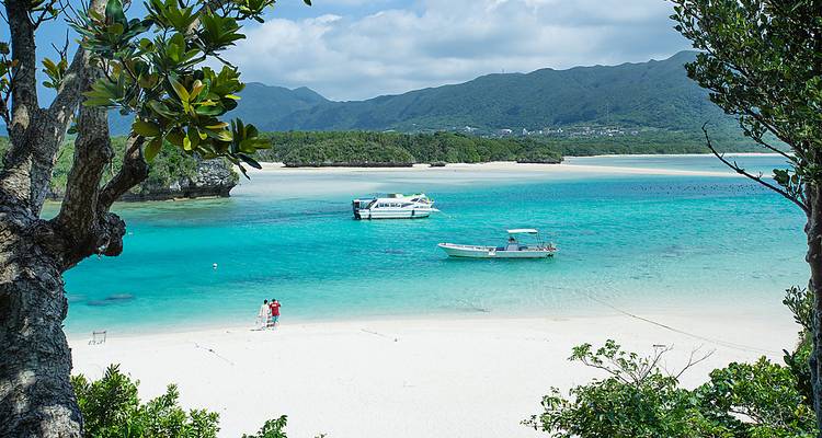 Unberührter Sandstrand mit weißem Sand, türkisblauem Wasser und verankerten Booten vor bewaldeten Bergen