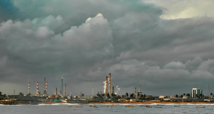 Complejo de refinería industrial en una costa brumosa bajo pesadas nubes grises con pequeños barcos de pesca en el mar