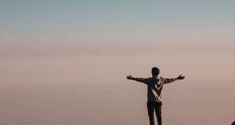 Un voyageur solitaire se tient les bras écartés sur un point de vue élevé au-dessus d'un océan de nuages sous un ciel aux teintes pastel.
