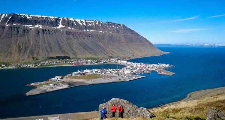 Vue depuis la colline surplombant la ville d'Ísafjörður et le fjord avec un plateau aux sommets enneigés sous un soleil éclatant.