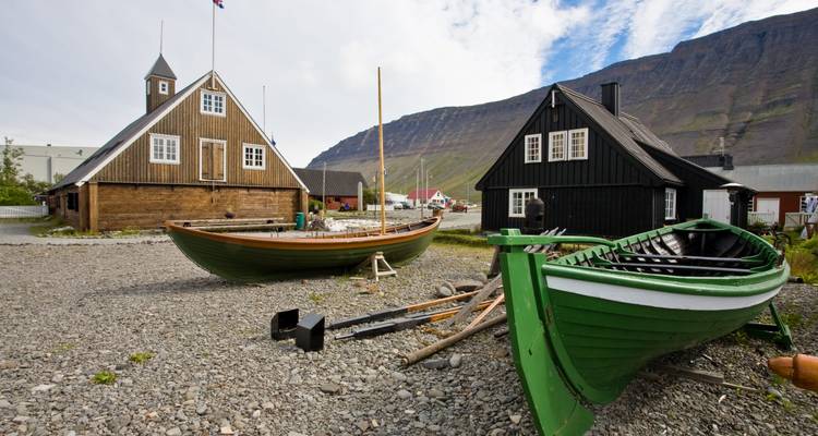 Bateaux de pêche traditionnels en bois sur gravier à côté de maisons aux toits de gazon avec une paroi de fjord abrupte en arrière-plan.