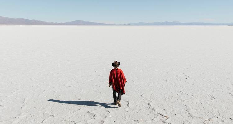 Una figura solitaria con un poncho rojo y un sombrero camina a grandes pasos por una llanura de sal blanca interminable.