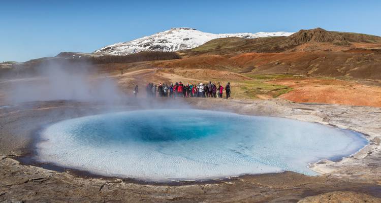 Bassin géothermique fumant avec un groupe de touristes observant en arrière-plan de montagnes enneigées.