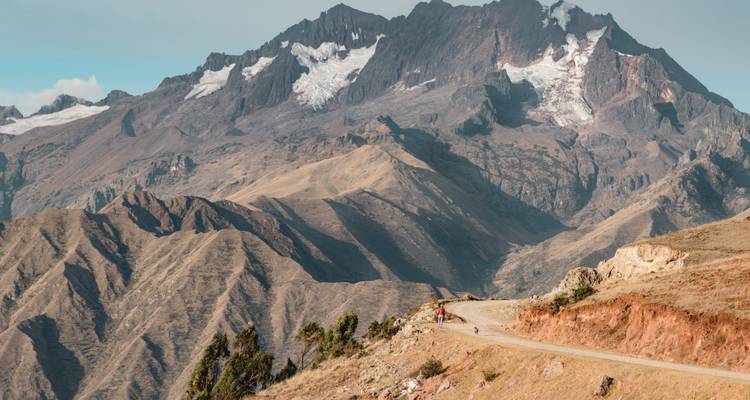 Amplio paisaje de montañas andinas con un camino de tierra sinuoso y un pico nevado bajo un cielo despejado.