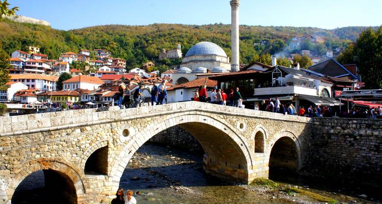 Stone footbridge over the Bistrica River with Prizren’s old bazaar, mosque, and hillside houses in the background