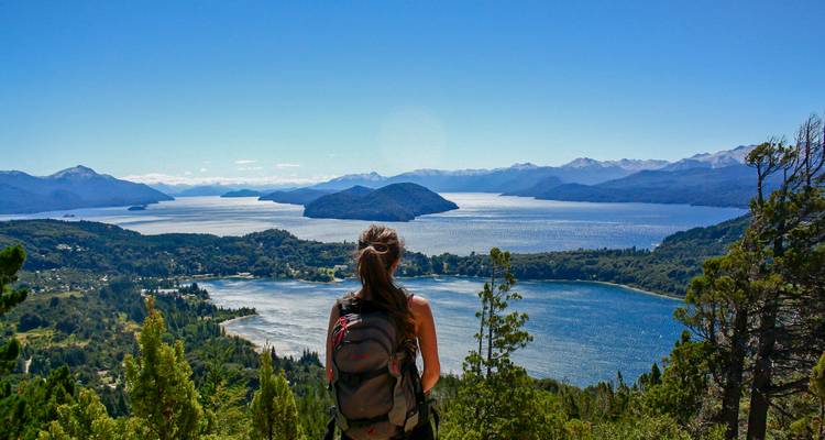 Ein Rucksacktourist blickt von einem Berggipfel über eine Seenkette und bewaldete Hügel.