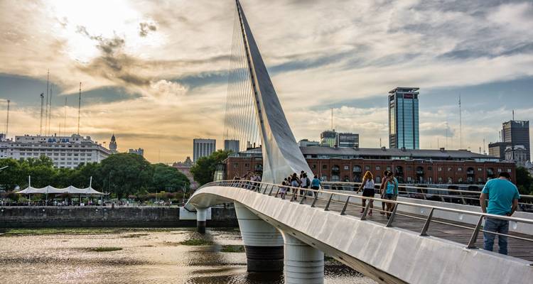 Fußgänger überqueren die moderne Puente de la Mujer-Brücke mit Stadtskyline im goldenen Abendlicht.