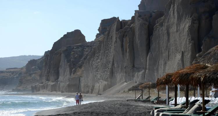 Des falaises volcaniques abruptes dominant une plage de sable noir où un couple se promène près de douces vagues.