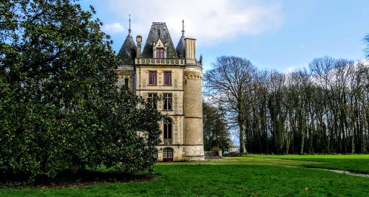 Petit château de pierre de conte de fées entouré d'une pelouse, d'un magnolia et d'une forêt dénudée.