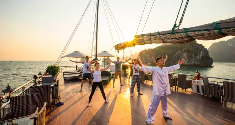 Passagiers oefenen zonsopgang Tai Chi op het houten dek van een cruiseschip met kalkstenen eilanden op de achtergrond.