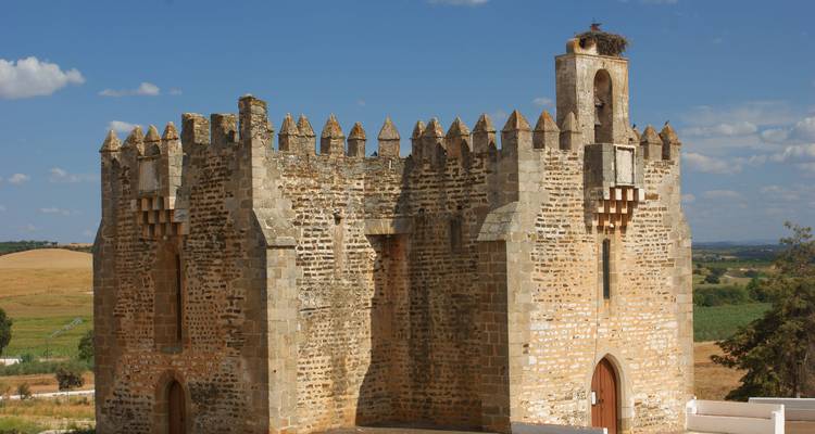 A fortified medieval tower with battlements set against rolling farmland under a clear sky.